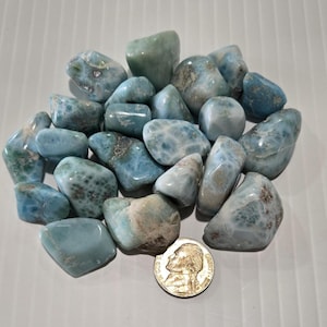 May include: A collection of polished larimar stones in shades of blue and white, with some showing brown inclusions. A US nickel coin is included for size reference. The stones vary in size and shape, with smooth, rounded surfaces.