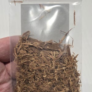 A hand holding a clear bag of shredded Cat's Claw bark, showing the natural orange-brown texture. This ethically sourced ritual herb is prepared by InSpyrit Herbs for spiritual grounding and protection.