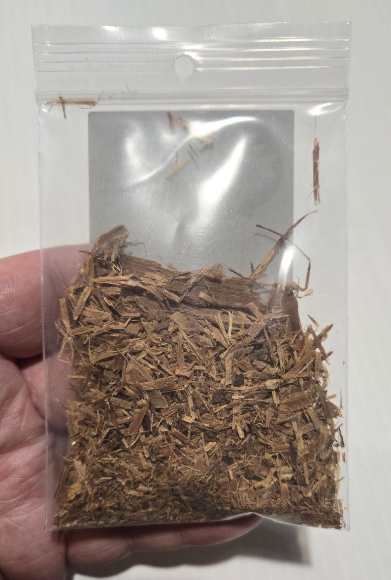 A hand holding a clear bag of shredded Cat's Claw bark, showing the natural orange-brown texture. This ethically sourced ritual herb is prepared by InSpyrit Herbs for spiritual grounding and protection.
