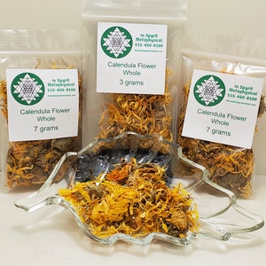 May include: Three clear plastic bags of dried calendula flowers, labeled "Calendula Flower Whole" with weights of 3 and 7 grams. A clear glass leaf-shaped dish holds more dried flowers. The text "In Spyrit Metaphysical" and a phone number are visible.