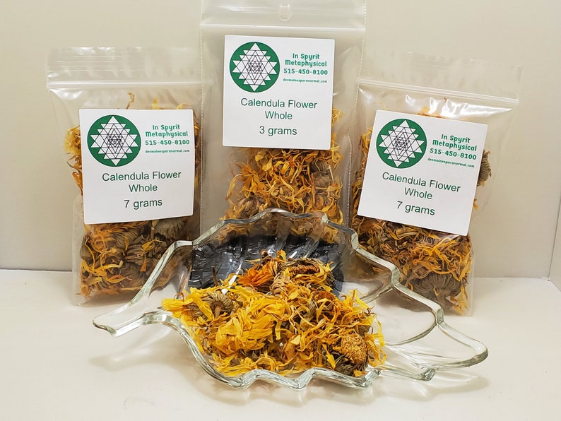 May include: Three clear plastic bags of dried calendula flowers, labeled "Calendula Flower Whole" with weights of 3 and 7 grams. A clear glass leaf-shaped dish holds more dried flowers. The text "In Spyrit Metaphysical" and a phone number are visible.