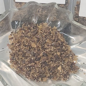 May include: A clear, leaf-shaped glass dish filled with dried, chopped seaweed. The seaweed is a mix of brown and tan pieces, with some lighter, granular elements. The dish sits on a white surface, and the overall presentation suggests a natural food product.
