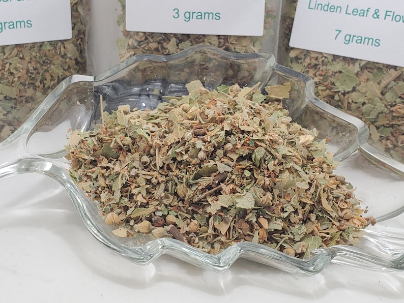 May include: Close-up of dried linden leaf and flower herbs in a clear glass dish. The herbs are a mix of green, brown, and tan. Small white labels with green text indicate the weight of the herbs in grams.