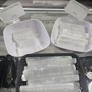 May include: The image displays several selenite crystal rods, some labeled as 2 inches and others as 4 inches. The crystals are displayed in white square bowls and a black basket. The text "Selenite 2 Inch Rods" and "Selenite 4 Inch Rods" is visible.