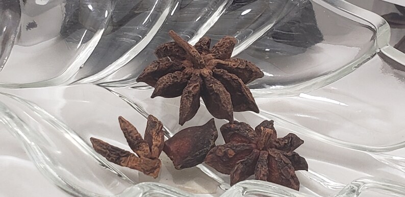 A close-up, high-angle view of several whole, dried Star Anise pods resting in a clear glass decorative dish. The star-shaped botanicals are a rich, earthy brown, showcasing their natural symmetry and texture, perfect for ritual use or culinary tea.