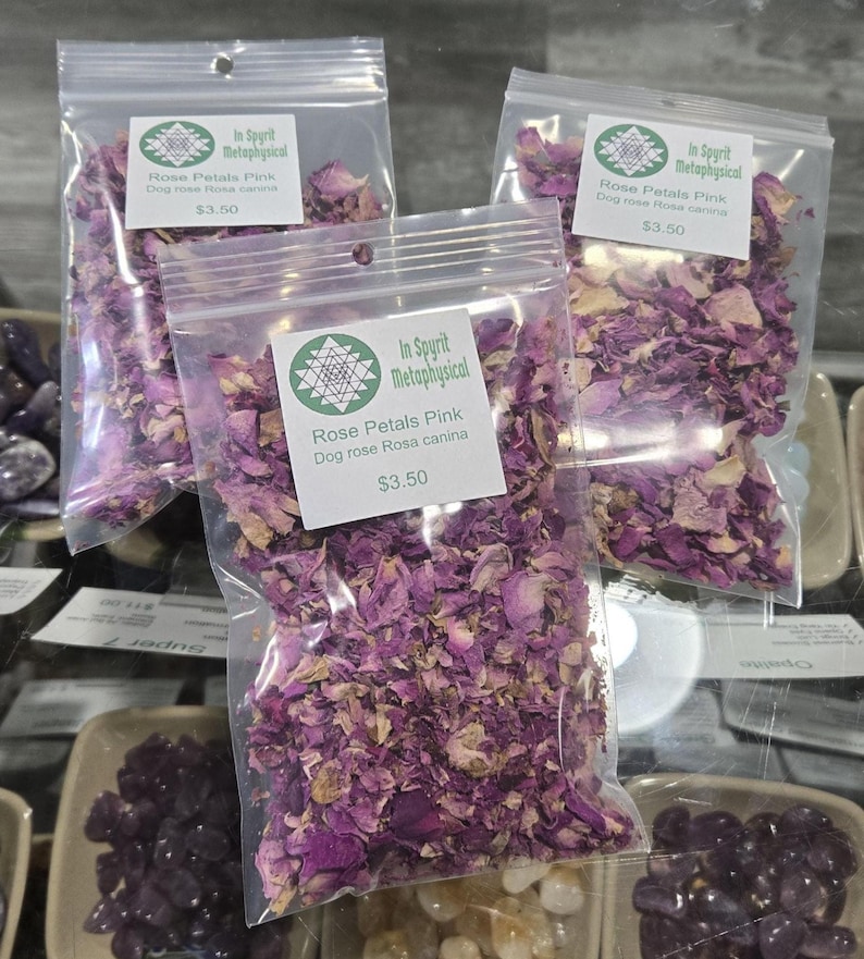 Clear botanical bag filled with dried pink rose petals by InSpyrit Herbs, showing a mix of vibrant fuchsia and soft pink floral textures against a natural background.