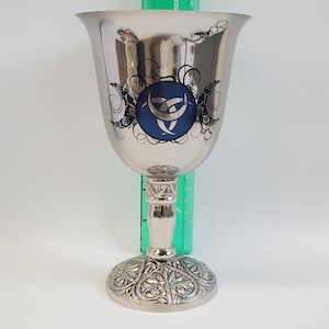 May include: Silver-colored chalice with a wide, flaring bowl and a detailed, ornate base. The bowl features a blue emblem with a crescent moon design, surrounded by black swirls. A green ruler is placed behind the chalice.