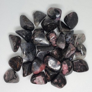 May include: A collection of tumbled stones, predominantly black with pink and gray inclusions. The stones are smooth and polished, with varying shapes and sizes. The overall aesthetic is natural and earthy, suitable for crystal enthusiasts or those interested in gemstones.