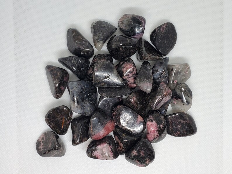May include: A collection of tumbled stones, predominantly black with pink and gray inclusions. The stones are smooth and polished, with varying shapes and sizes. The overall aesthetic is natural and earthy, suitable for crystal enthusiasts or those interested in gemstones.