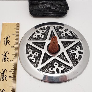 May include: A silver incense burner with a pentagram design, holding a brown incense cone. A black crystal and a wooden ruler are also present. The incense burner is approximately 10 cm in diameter.