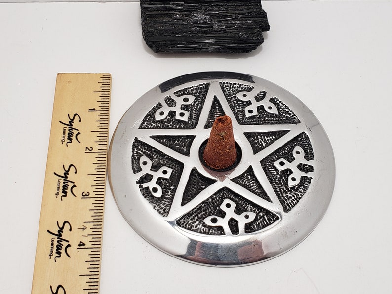 May include: A silver incense burner with a pentagram design, holding a brown incense cone. A black crystal and a wooden ruler are also present. The incense burner is approximately 10 cm in diameter.