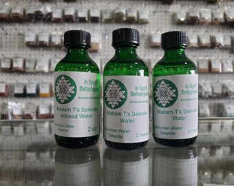May include: Three green glass bottles with black caps, labeled "Madam T's Selenite Infused Water" and "Madam T's Selenite Water." Each bottle has a white label with a geometric design and the text "In Spirit Metaphysical." The bottles are 2 oz each.