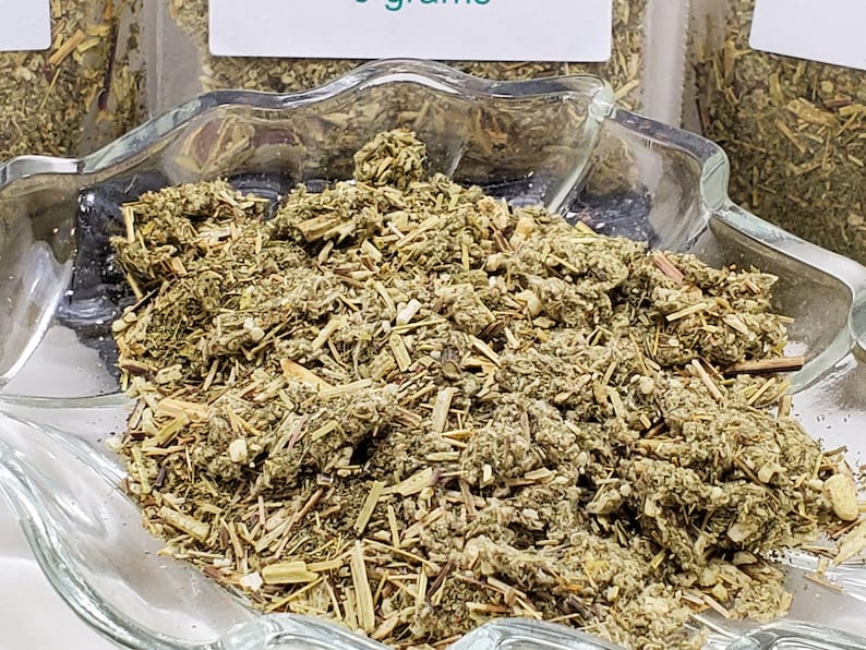 High-angle view of loose dried Mugwort (Artemisia vulgaris) displayed in a clear glass leaf-shaped ritual dish. The silver-green botanical texture is clearly visible against the white background, showcasing the quality of InSpyrit Metaphysical ritual herbs.