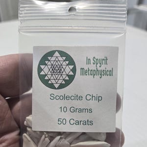 A close-up of a small, clear resealable bag filled with white Scolecite chip stones. The label is clearly visible with the InSpyrit Metaphysical logo and the text "Scolecite Chip 10 Grams 50 Carats