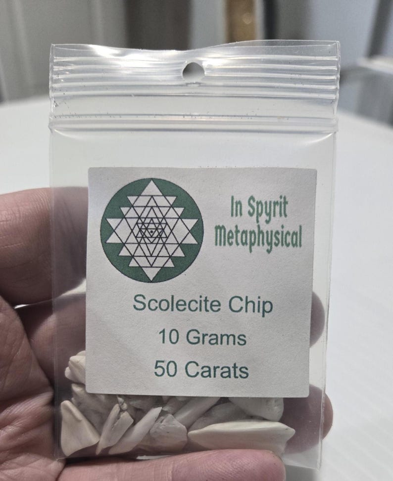 A close-up of a small, clear resealable bag filled with white Scolecite chip stones. The label is clearly visible with the InSpyrit Metaphysical logo and the text "Scolecite Chip 10 Grams 50 Carats