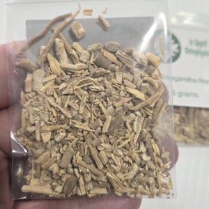 A hand holding a clear bag of dried Ashwagandha Root from In Spryit Metaphysical, showing the natural tan color and shredded texture of the botanical against a neutral background.