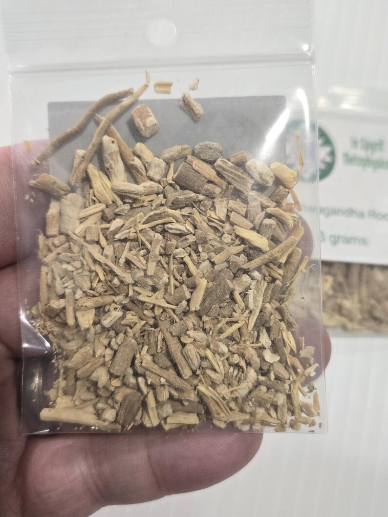 A hand holding a clear bag of dried Ashwagandha Root from In Spryit Metaphysical, showing the natural tan color and shredded texture of the botanical against a neutral background.