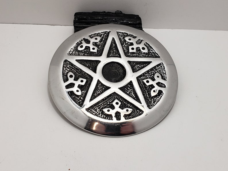 May include: A silver-coloured, circular pendant featuring a raised pentagram design. The pentagram is centred with a black circle and surrounded by decorative patterns. The pendant is likely made of metal and has a polished finish.