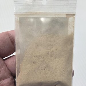 A hand holding a clear bag of finely ground Ashwagandha Root Powder, showing the natural tan color and sandy texture of the Withania Somnifera herb. This close-up provides a clear view of the powder's consistency for use in wellness lattes or ritual work.