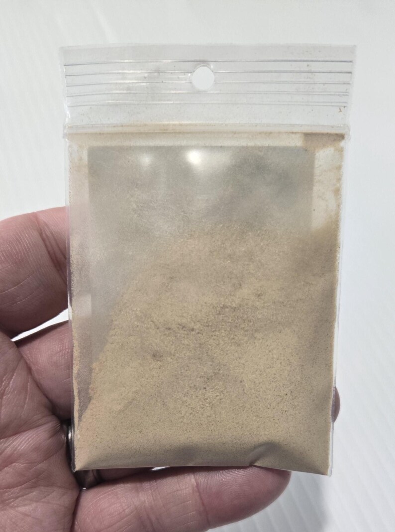 A hand holding a clear bag of finely ground Ashwagandha Root Powder, showing the natural tan color and sandy texture of the Withania Somnifera herb. This close-up provides a clear view of the powder's consistency for use in wellness lattes or ritual work.