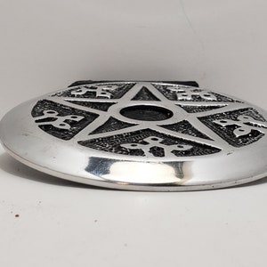 May include: Silver oval belt buckle featuring a raised pentagram design. The pentagram is centrally placed, surrounded by decorative patterns. The buckle has a shiny, metallic finish and a black leather backing.
