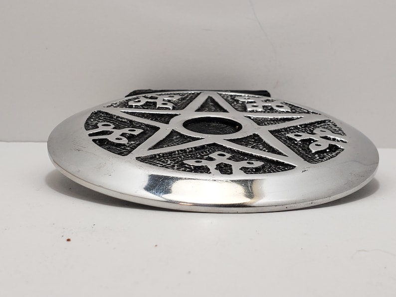 May include: Silver oval belt buckle featuring a raised pentagram design. The pentagram is centrally placed, surrounded by decorative patterns. The buckle has a shiny, metallic finish and a black leather backing.