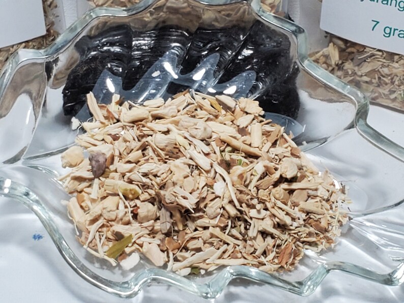A close-up photograph by InSpyrit Metaphysical showing dried, cut hydrangea root pieces piled neatly inside a clear, textured glass leaf-shaped dish.