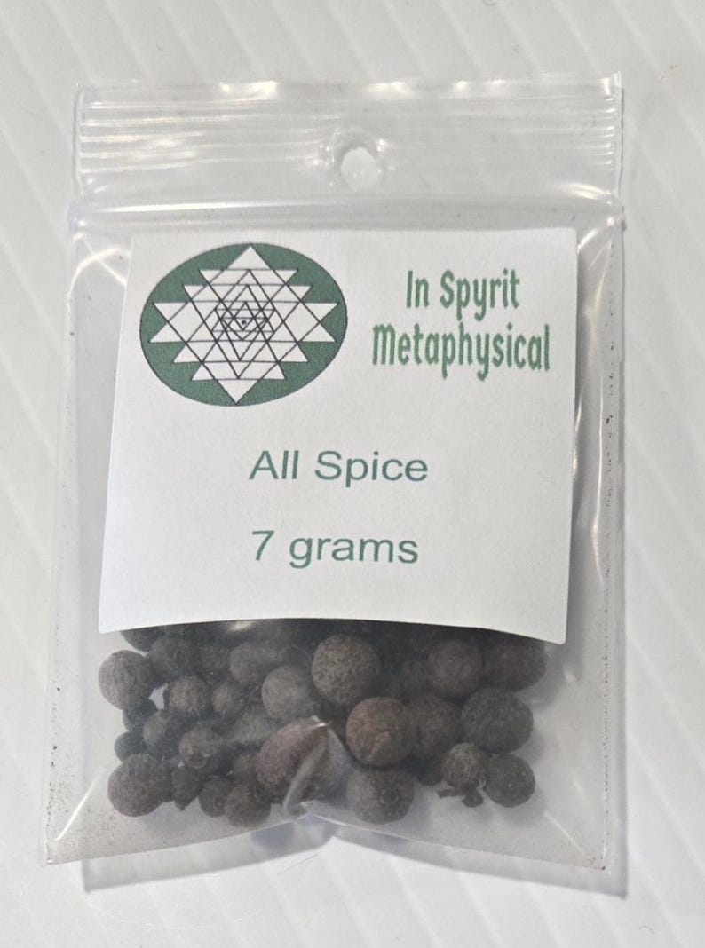 A small, clear plastic bag containing 7 grams of whole Allspice berries from InSpryitMetaphysical. The white label at the top displays the green and black shop logo, the product name "All Spice," and the "7 grams" weight. The dark brown, dried berries are visible through the transparent packaging against a light gray background.