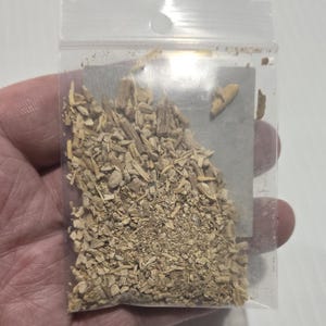 A hand holding a clear bag of dried Ashwagandha Root from In Spryit Metaphysical, showing the natural tan color and shredded texture of the botanical against a neutral background.
