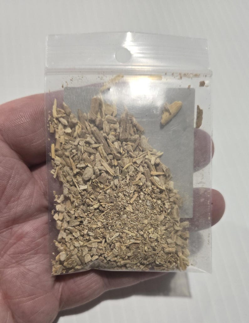 A hand holding a clear bag of dried Ashwagandha Root from In Spryit Metaphysical, showing the natural tan color and shredded texture of the botanical against a neutral background.