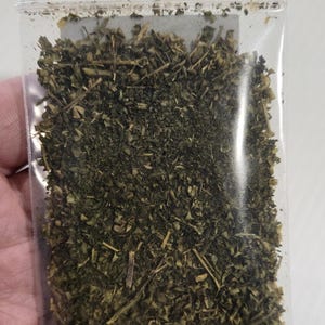 A hand holding a 7-gram clear ritual bag filled with dried organic Skullcap Leaf from In Spyrit Metaphysical, showcasing the texture and quality of the earthy botanical blend inside.