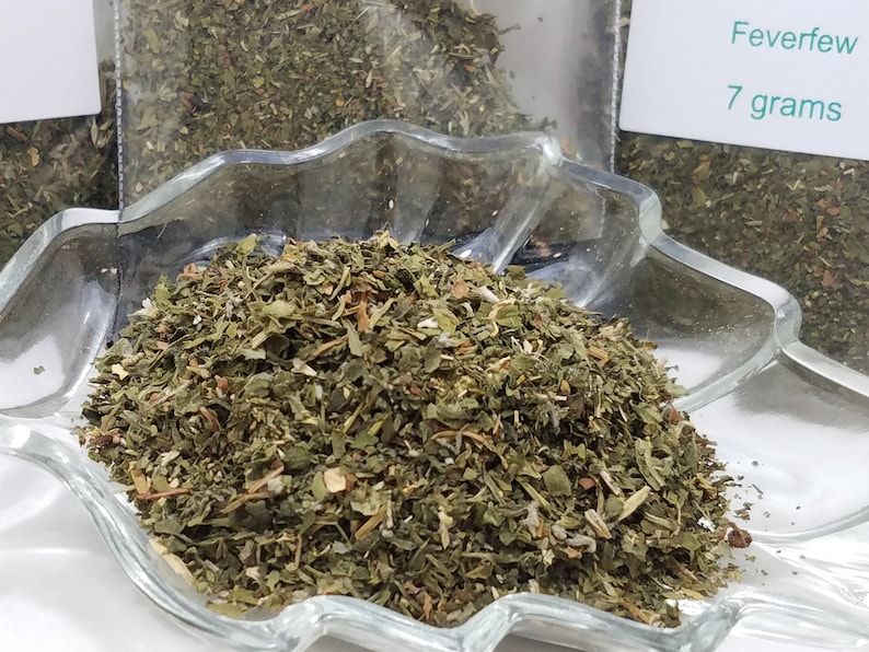 May include: A clear glass dish filled with dried Feverfew herbs. The herbs are a mix of green and brown, with a leafy texture. A label reads "Feverfew 7 grams".