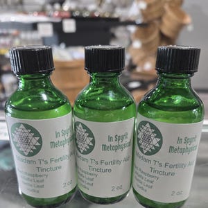 Könnte beinhalten: Drei grüne Glasflaschen mit schwarzen Verschlüssen. Jede Flasche hat ein weißes Etikett mit dem Text "In Spyrit Metaphysical Madam T's Fertility Aid Tincture". Die Etiketten listen auch die Zutaten auf, und die Flaschen sind je 59 ml groß.
