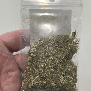 A hand holding a clear, unlabeled bag of dried Mugwort herb against a white background. The photo shows a close-up of the natural, shredded texture of the silver-green Artemisia vulgaris leaves, providing a clear view of the product's quality.