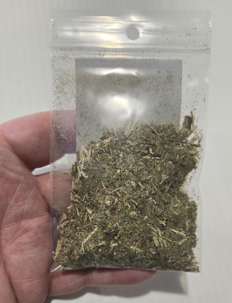 A hand holding a clear, unlabeled bag of dried Mugwort herb against a white background. The photo shows a close-up of the natural, shredded texture of the silver-green Artemisia vulgaris leaves, providing a clear view of the product's quality.