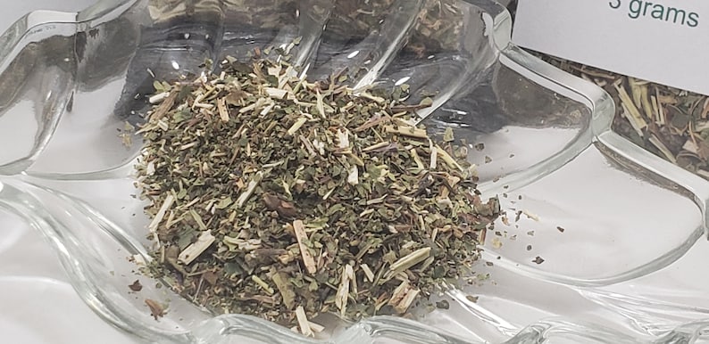 Dried organic Skullcap Leaf from In Spyrit Metaphysical, displayed in a clear, leaf-shaped bowl. The natural, earthy green botanical blend is showcased with several small, clear packaging bags arranged in the background.