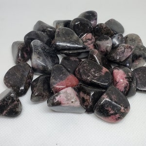 May include: A pile of tumbled rhodonite stones. The stones are a mix of dark gray and black, with patches of pink and red. The stones are smooth and polished, reflecting light. The overall theme is natural gemstones.