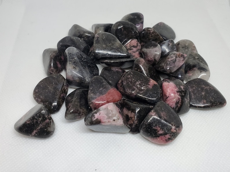 May include: A pile of tumbled rhodonite stones. The stones are a mix of dark gray and black, with patches of pink and red. The stones are smooth and polished, reflecting light. The overall theme is natural gemstones.