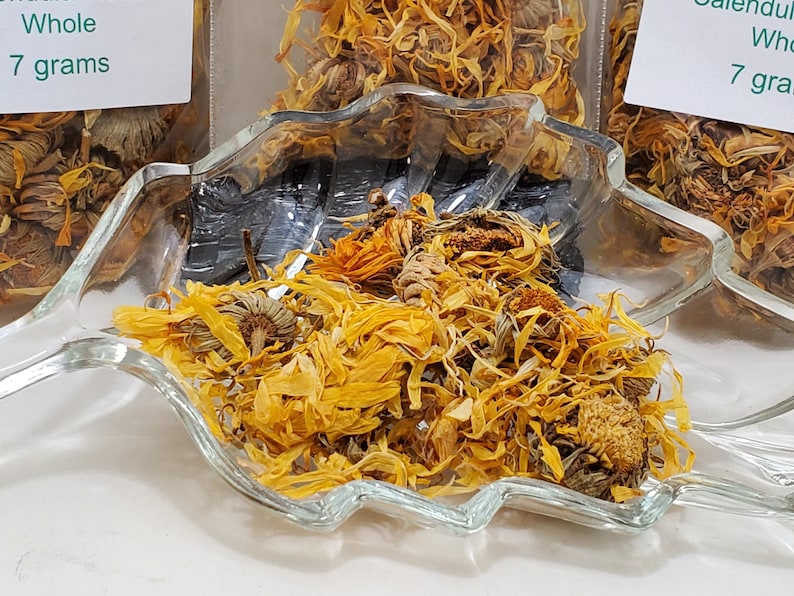 May include: Dried calendula flowers in a clear glass leaf-shaped dish. The flowers are a vibrant yellow and orange, with some whole flower heads visible. The image also shows bags of calendula flowers with labels that read "Whole 7 grams".