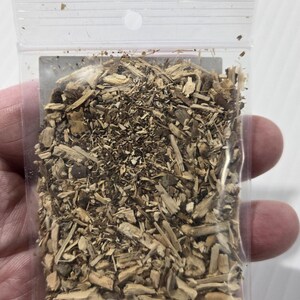 A photograph from InSpyrit Metaphysical showing a hand holding up a small, clear plastic bag filled with dried hydrangea root to display the scale of the product and its natural botanical texture.