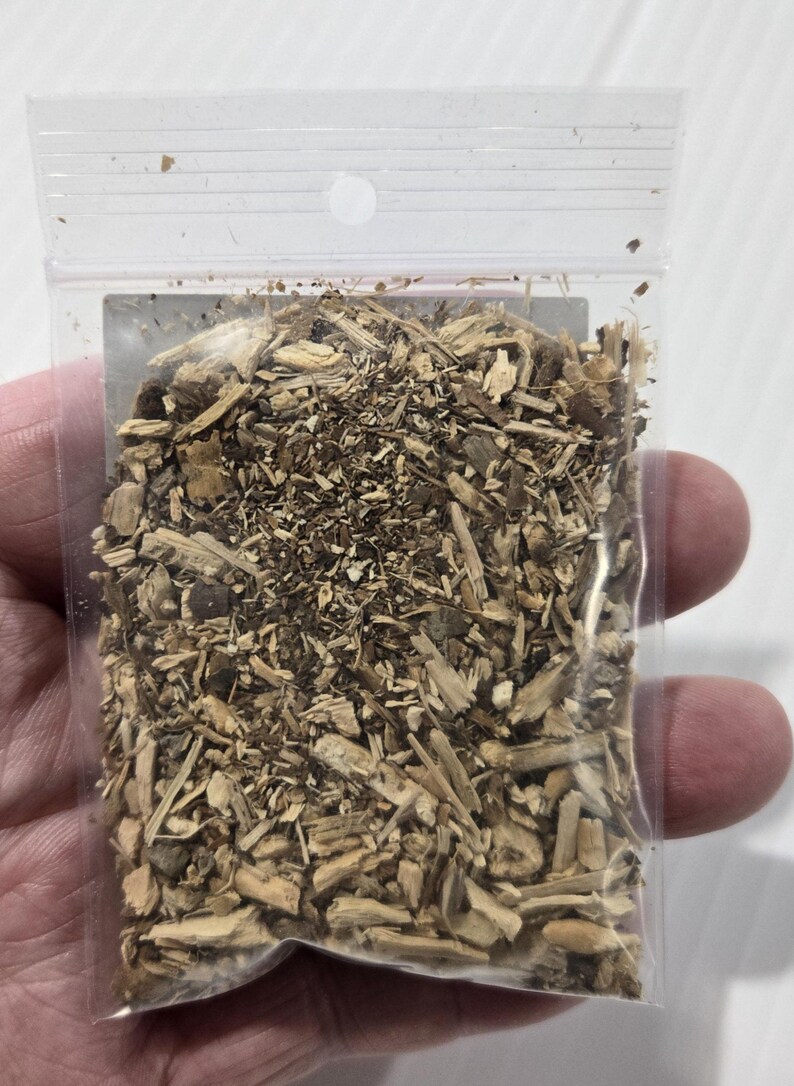 A photograph from InSpyrit Metaphysical showing a hand holding up a small, clear plastic bag filled with dried hydrangea root to display the scale of the product and its natural botanical texture.