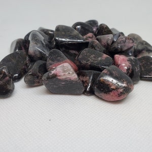 May include: A pile of tumbled rhodonite stones. The stones are a mix of black and pink, with a polished, glossy finish. The pink hues vary in intensity, creating a visually appealing contrast. These stones are often used for their purported healing properties.