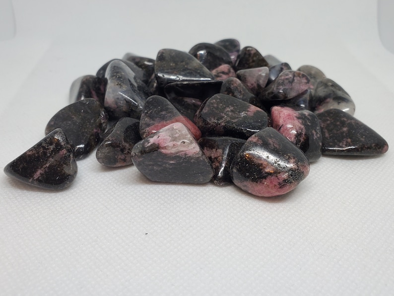 May include: A pile of tumbled rhodonite stones. The stones are a mix of black and pink, with a polished, glossy finish. The pink hues vary in intensity, creating a visually appealing contrast. These stones are often used for their purported healing properties.
