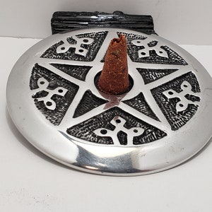 May include: A silver incense holder featuring a pentagram design and a brown incense cone. The holder is circular with decorative patterns around the pentagram. A black stone is behind the holder.