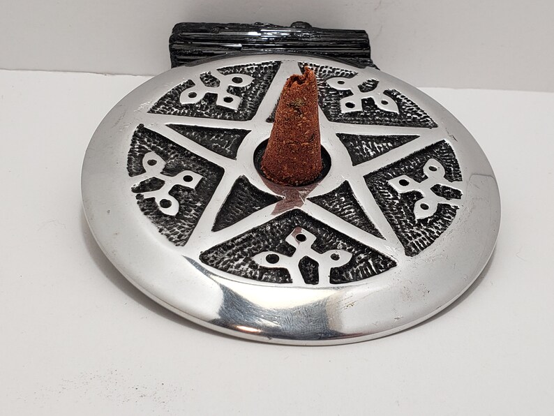May include: A silver incense holder featuring a pentagram design and a brown incense cone. The holder is circular with decorative patterns around the pentagram. A black stone is behind the holder.