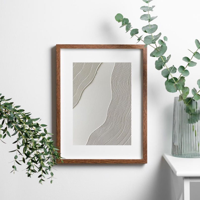 May include: A framed abstract art print featuring a minimalist design with flowing, textured lines in shades of gray and white. The artwork is encased in a warm brown wooden frame, creating a modern and elegant aesthetic. The print is displayed on a white wall.