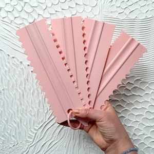 May include: A set of six pink plastic cake scrapers with various edge designs. Each scraper has a different scalloped or ridged edge for creating decorative patterns on cakes and pastries. A small ring is at the end of each scraper for hanging.