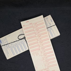 May include: A stack of white time cards with red and black print. The cards have fields for name, number, date, and time in and out. The cards are used for tracking employee hours.