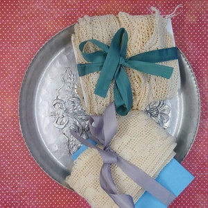 May include: Two pieces of white crocheted fabric, one tied with a teal ribbon and the other with a gray ribbon, are displayed on a silver tray with an ornate design. The tray is on a red background with white polka dots.