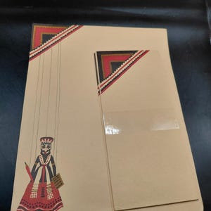 May include: Tan stationery set featuring a puppet illustration with red, black, and white accents. The puppet holds a small sign. The set includes a large sheet and a smaller card, both with decorative corner designs.